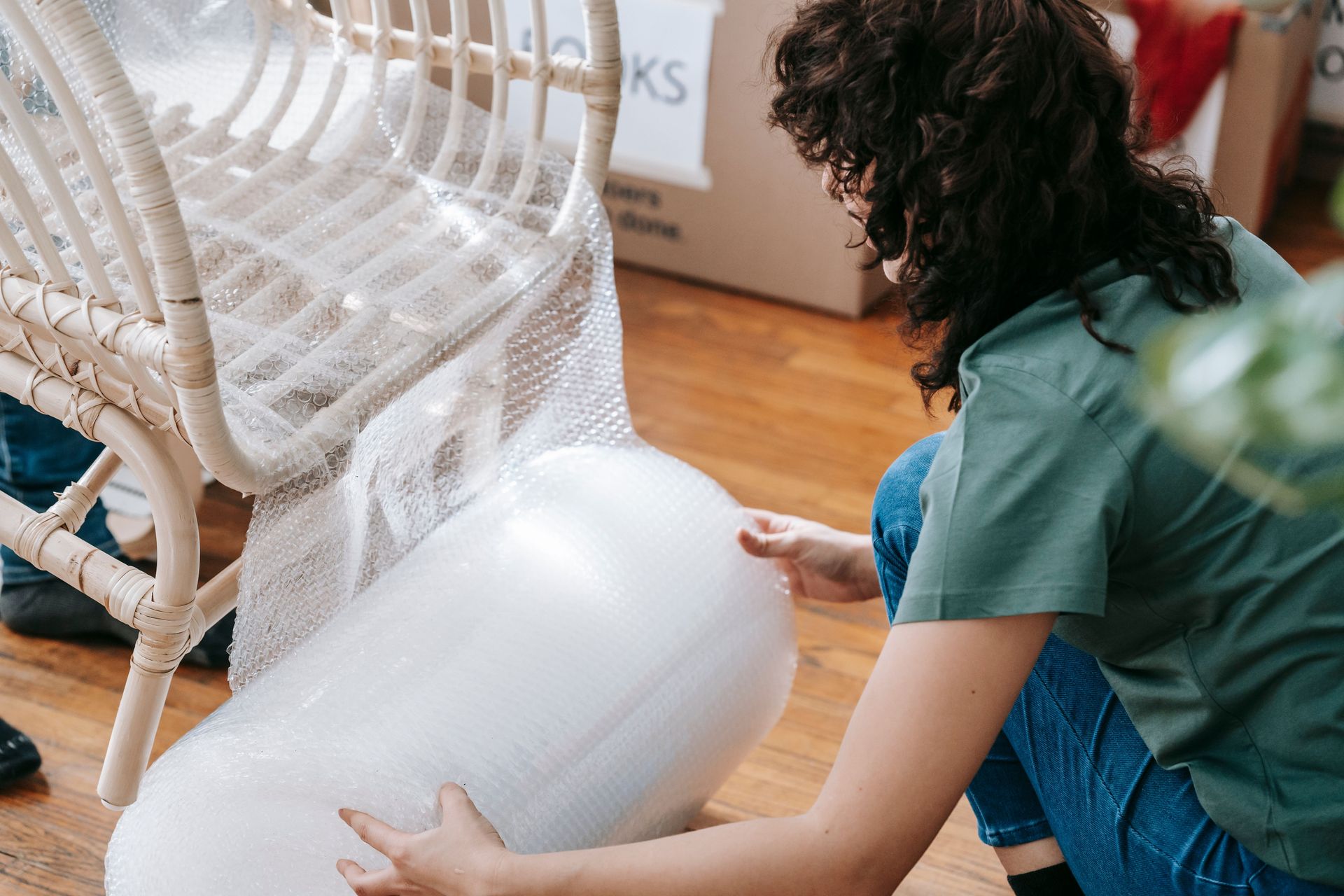 Woman kneeling to wrap a wicker chair with bubble wrap, preparing for relocation.